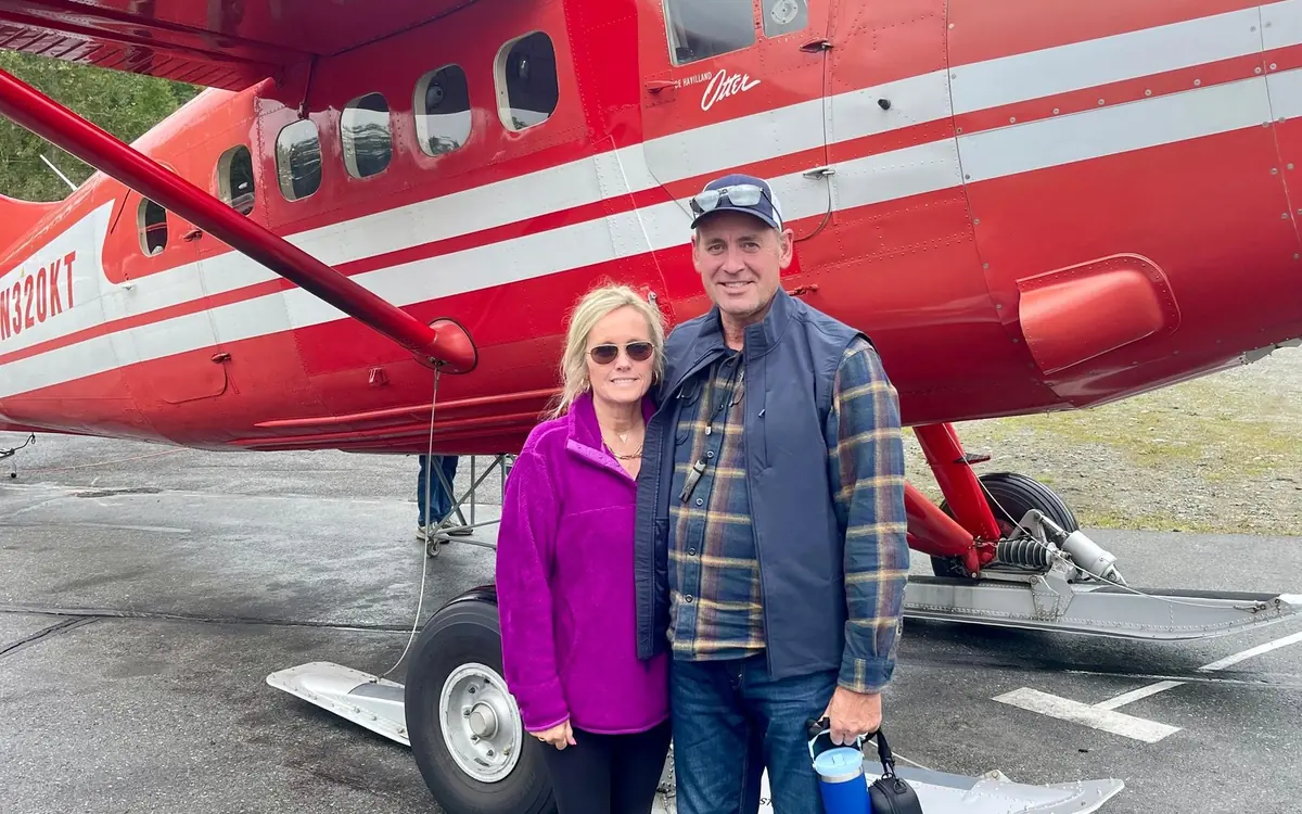 Passengers standing on glacier snow beside a red ski plane with the peaks of the Alaska Range behind them