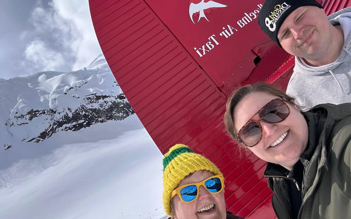 Three passengers standing on the snow beside the red tail of a ski plane