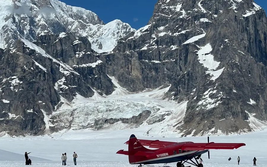 A red ski plane on a glacier with passengers on the snow below a wall of rock and ice