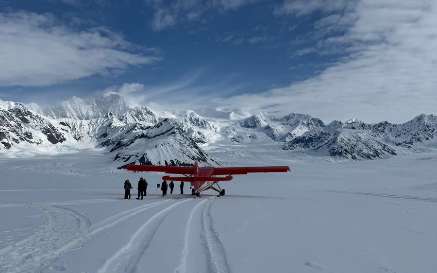 A red ski plane parked on a wide snowfield with passengers beside it and the peaks of the Alaska Range behind