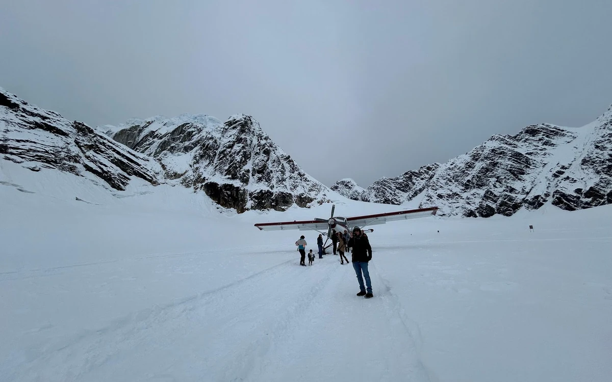 A person walking on snow beside a parked ski plane with peaks behind